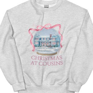 Peut inclure: Sweat-shirt gris clair avec un motif de boule à neige représentant une maison avec des accents roses et un ruban rose. Le texte "CHRISTMAS AT COUSINS" est imprimé en dessous.