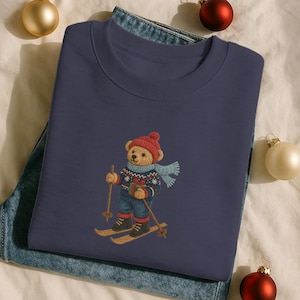 Könnte beinhalten: Marineblaues Sweatshirt mit einem gestickten Bären, der eine rote Mütze, einen blauen Schal und eine blaue Jeans trägt und Ski fährt. Der Bär hält Skistöcke und ein Geschenk. Das Sweatshirt ist auf einer Jeans gefaltet. Weihnachtskugeln umgeben das Sweatshirt.