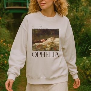 May include: Cream-coloured sweatshirt displaying the painting "Ophelia" by John Everett Millais. The artwork shows a woman in a white dress in water, with the word "Ophelia" printed below. The sweatshirt is worn by a person in a garden setting.