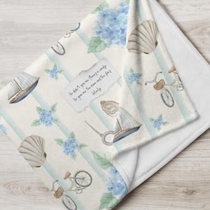 May include: A soft, white throw blanket with a pattern of blue hydrangeas, seashells, bicycles, and sailboats on a striped background. A small piece of paper with text is also visible.