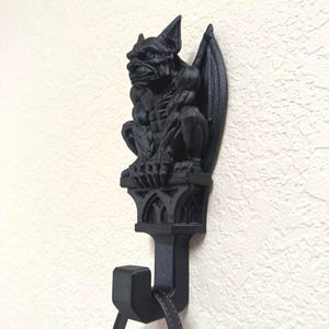 Pode incluir: Um gancho de parede preto fosco com uma figura de gárgula esculpida pousada sobre uma base arquitetônica de estilo gótico. A gárgula tem orelhas pontudas, asas dobradas e um corpo musculoso. Um gancho estende-se para baixo a partir da base.