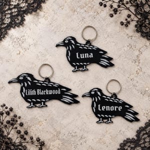 Pode incluir: Três porta-chaves em forma de corvo preto com detalhes gravados a branco e argolas de metal prateado. Cada porta-chaves apresenta um nome gravado em letra gótica: Luna, Lilith Blackwood e Lenore. As silhuetas dos corvos são planas e detalhadas.