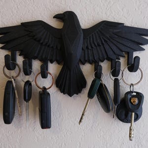 May include: Black raven-shaped key holder with outstretched wings, featuring multiple hooks for keys. The key holder is mounted on a wall and holds various car keys and keyrings. The raven design adds a decorative touch to any hallway.
