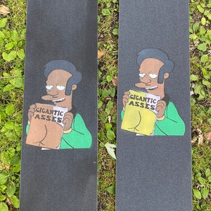 May include: Two black skateboard decks with a cartoon character holding a magazine with the text "Gigantic Asses". The character is wearing a green shirt and has black hair.