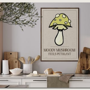 Peut inclure: Une affiche encadrée avec une illustration de champignon fantaisiste, chapeau jaune, et le texte "Moody Mushroom Feels Petulant". L'affiche est dans une cuisine avec des planches et ustensiles en bois.