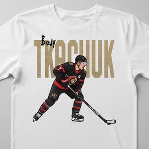 May include: White t-shirt featuring a hockey player in a black and red uniform with the name "Brady Tkachuk" in gold block letters. The player is in a skating pose, holding a hockey stick.