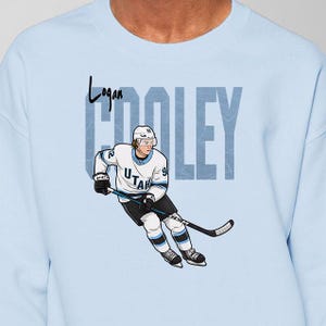 May include: Pale blue sweatshirt with a cartoon hockey player in a white and blue uniform. The name "Logan" is above the player, and the name "COOLEY" is behind the player in a blocky font.
