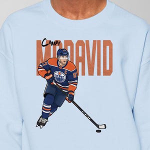 May include: Pale blue sweatshirt with a cartoon image of a hockey player in blue and orange. The player is holding a hockey stick, and the name "Connor McDavid" is printed above the player in a large font.