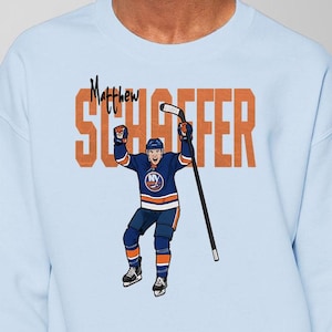 May include: Pale blue sweatshirt with a cartoon hockey player in a blue and orange uniform, celebrating with raised arms. The name "Matthew SCHAFFFER" is printed above the player in orange block letters.