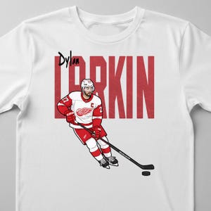 May include: White t-shirt featuring a graphic of a hockey player in a red and white uniform, with the name "Dylan Larkin" above and "LARKIN" in large red letters. The player is holding a hockey stick and puck.