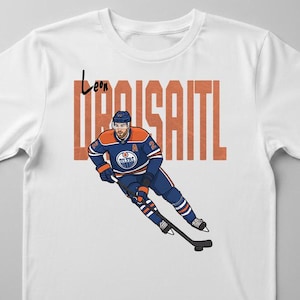 Könnte beinhalten: Weißes T-Shirt mit einer Cartoon-Illustration eines Eishockeyspielers in blau-oranger Uniform. Der Name "Leon Draisaitl" ist in orangefarbenen Blockbuchstaben aufgedruckt. Der Spieler hält einen Hockeyschläger.