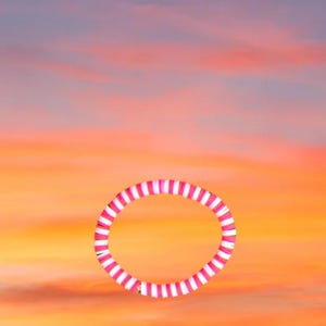 May include: A pink and white striped bracelet against a vibrant sunset sky. The bracelet is a circular shape, with alternating pink and white stripes. The background features a gradient of orange, pink, and blue hues.