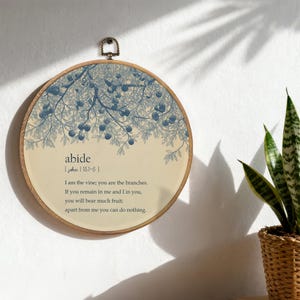 Pode incluir: Arte de parede redonda em madeira com uma ilustração botânica azul e creme. O design inclui um motivo de galho de árvore e a palavra "abide" com um versículo bíblico. Possui um laço de metal para pendurar.