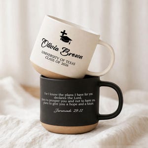 May include: Two ceramic mugs, one stacked on top of the other. The top mug is white with a black graduation cap and cross graphic, and the name "Olivia Brown" and "UNIVERSITY OF TEXAS CLASS OF 2026" printed on it. The bottom mug is black with a Bible verse printed on it.