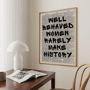 May include: A framed print featuring the quote "WELL BEHAVED WOMEN RARELY MAKE HISTORY" in black, bold lettering. The background is a collage of dictionary pages. The frame is light brown.