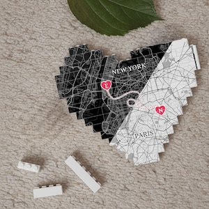 May include: A heart-shaped map of New York and Paris, with a dotted line connecting the cities. The map is black and white, with the cities labeled. Two small white building blocks are in the foreground.