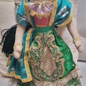 May include: A handmade doll dressed in a traditional Indian outfit. The doll has a painted face with detailed features, a gold crown, and wears a green and gold embroidered dress. The doll is seated on a light-colored couch.