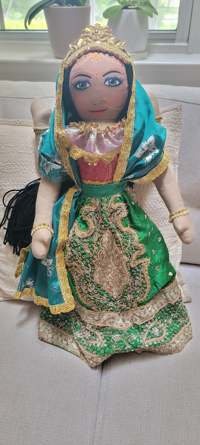 May include: A handmade doll dressed in a traditional Indian outfit. The doll has a painted face with detailed features, a gold crown, and wears a green and gold embroidered dress. The doll is seated on a light-colored couch.
