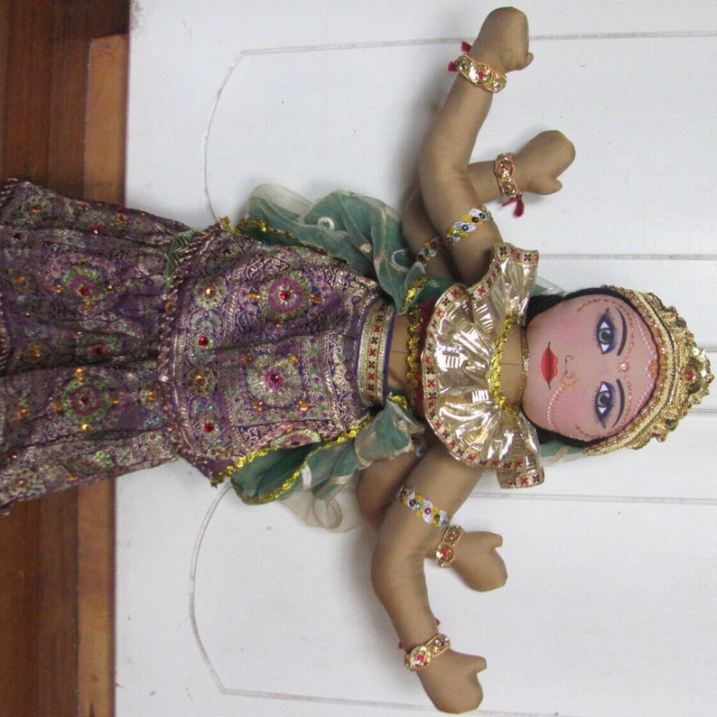 May include: A fabric doll dressed in a traditional Indian outfit. The doll has a painted face with large blue eyes, a red nose ring, and a gold headdress. The doll's outfit is purple and green with gold accents and red and gold embellishments.
