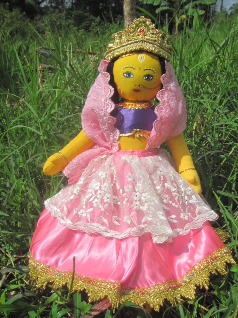 May include: A handmade doll with a yellow face, blue eyes, and a gold crown. The doll wears a pink and white lace skirt, a purple top, and a pink veil. The skirt is trimmed with gold and colorful beads. The doll is set against a backdrop of green grass.