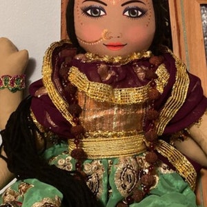 May include: A handcrafted doll with a serene expression, adorned with a gold crown and intricate makeup. The doll wears a maroon and gold embellished outfit with a green skirt. A beaded bracelet is visible on the left arm. The doll's hair is black.