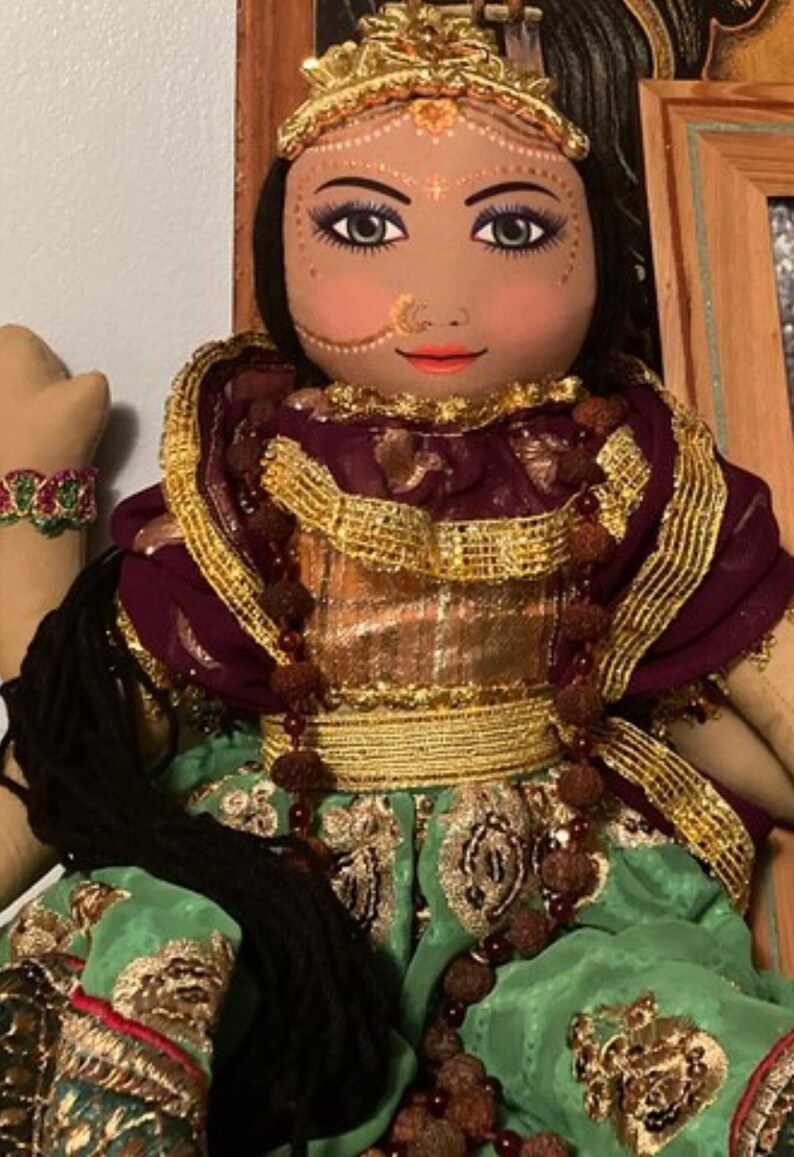 May include: A handcrafted doll with a serene expression, adorned with a gold crown and intricate makeup. The doll wears a maroon and gold embellished outfit with a green skirt. A beaded bracelet is visible on the left arm. The doll's hair is black.