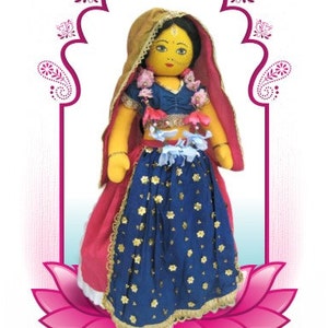 May include: A handmade doll dressed in traditional Indian attire. The doll wears a blue skirt with gold floral accents, a red shawl, and a gold head covering. The doll is set against a pink lotus flower and an ornate arch.
