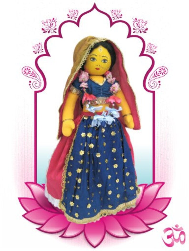 May include: A handmade doll dressed in traditional Indian attire. The doll wears a blue skirt with gold floral accents, a red shawl, and a gold head covering. The doll is set against a pink lotus flower and an ornate arch.