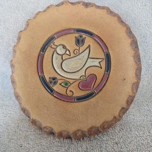 May include: A round, ceramic coaster with a decorative bird design. The bird is white with a black outline, surrounded by a ring of blue, pink, and green accents. The coaster has a textured, light brown surface and a scalloped edge.