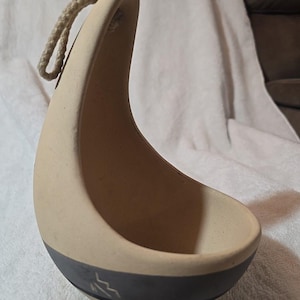 May include: A beige ceramic hanging planter with a dark gray band around the base. The planter has a curved handle with a rope for hanging. The planter's design is minimalist and modern, suitable for small plants or decorative items.