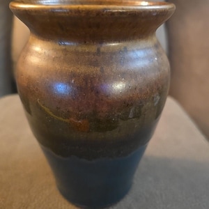 May include: A small, handcrafted ceramic vase with a unique color gradient. The top rim is a warm brown, transitioning to a bronze and purple midsection, and finishing with a deep blue base. The vase has a slightly flared top and a rounded body.