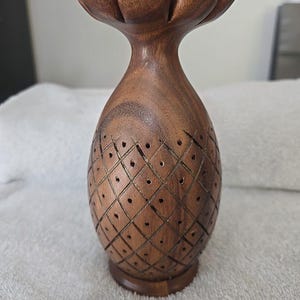 May include: A handcrafted wooden pineapple-shaped decorative item. The top features a flower-like design, while the body has a diamond pattern with small holes. The item is made of a rich brown wood and stands on a round base.