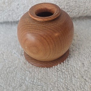 May include: A small, round wooden vase with a wide opening and a circular base. The vase is made of light brown wood and has a smooth, polished finish. The vase is a decorative item.