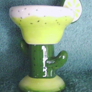May include: A ceramic margarita glass shaped like a cactus. The glass has a white rim with black speckles, a light green body, and a green cactus stem. A lime slice is attached to the rim.