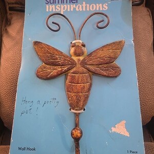 May include: A decorative wall hook shaped like a bee, with brown wings and body, and a yellow head. The hook is attached to a blue card with the text "summer inspirations" and "Hang a pretty pot!"