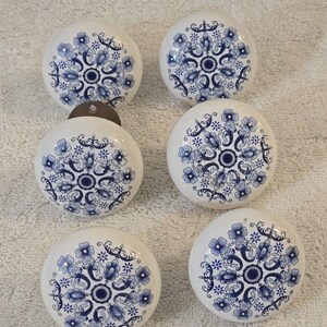 May include: Six white ceramic cabinet knobs with a blue floral design. The knobs are round with a metal base and feature a detailed pattern of flowers and leaves. The blue and white color scheme gives a classic, vintage feel.