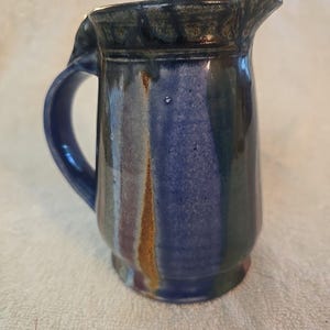 May include: A handmade ceramic pitcher with a dark blue handle and spout. The pitcher has a blue, green, and brown color scheme with a glossy finish. The pitcher is a decorative item for the kitchen.