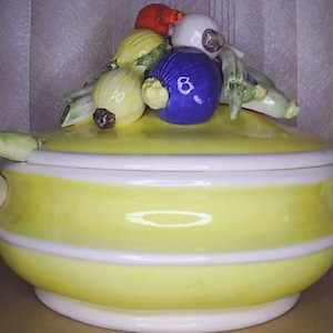 May include: A yellow ceramic serving dish with a lid, adorned with white stripes and handles. The lid is topped with ceramic vegetables, including red, white, yellow, and blue onions and green stalks. A decorative kitchenware item.