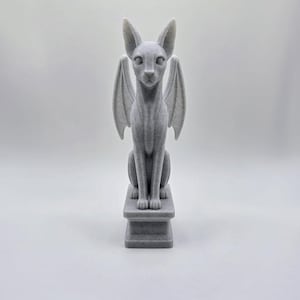 Puede incluir: Una figura impresa en 3D de color gris claro y textura rugosa que representa un gato sentado con alas de murciélago, sobre un pedestal rectangular. La base de la figura presenta el texto en relieve TinkerLayer en gris oscuro sobre el fondo.