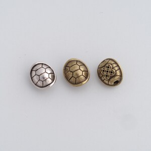 May include: Three metal turtle-shaped beads in silver, gold, and bronze. Each bead has a textured shell design. The bronze bead features a four-leaf clover design. These beads are ideal for jewelry making.