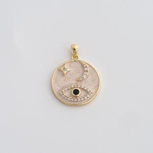 May include: Gold-tone pendant with a circular design featuring an eye, crescent moon, and star. The eye and moon are embellished with small, clear crystals. The background has a cream-colored, marbled effect.