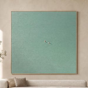 May include: A large square wall art piece featuring a solid, textured sage green surface with two tiny, distinct figures positioned near the center. The artwork is framed in a thin, light-colored wood border and hangs centered above a long sofa.
