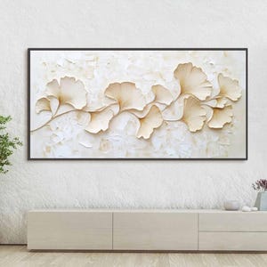 May include: A framed wall art piece featuring a relief of stylized ginkgo leaves in shades of cream and gold. The artwork is set against a textured, off-white background and framed in black. A decorative piece for home decor.