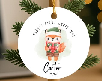 May include: A round white Christmas ornament featuring a fox illustration, wearing a hat and holding a gift. The text reads "Baby's First Christmas", with the name "Carter" and the year "2025". The ornament is suspended from a small wooden tree.