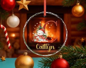 May include: A clear glass Christmas ornament with a red ribbon. The ornament features a snowman, deer, Christmas tree, and the name "Caitlyn" in gold script. A large red letter "C" is also present.