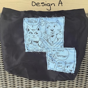 May include: Black tote bag with two light blue patches featuring cat illustrations and speech bubbles. One patch says "MEOW" and "Ya Totally Girl". The other says "WE SHOULD GO THRIFTING!" and "Nice Idea". The text "Design A" is above the bag.