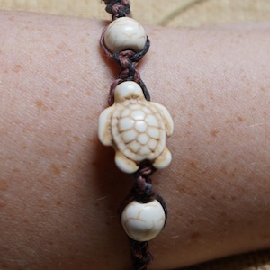 May include: A handmade bracelet featuring a turtle charm and two round beads. The turtle charm and beads are off-white, and the bracelet is made with a brown and black woven cord. The bracelet is shown on a person's arm.