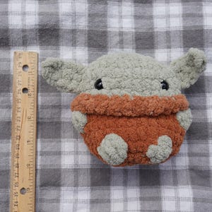 以下が含まれることがあります： 大きな耳を持つキャラクターの形をした手作りのぬいぐるみ。おもちゃは主にグレーで、オレンジブラウンの衣服を着ています。おもちゃの高さは約15cmです。