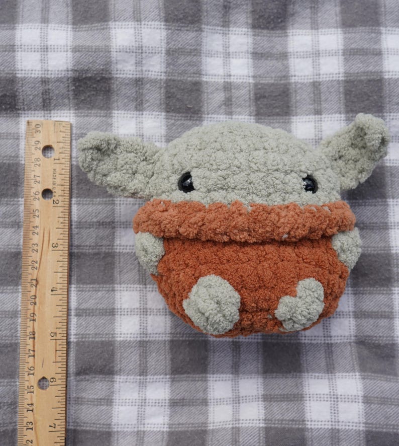 以下が含まれることがあります： 大きな耳を持つキャラクターの形をした手作りのぬいぐるみ。おもちゃは主にグレーで、オレンジブラウンの衣服を着ています。おもちゃの高さは約15cmです。