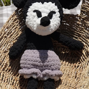 May include: A handmade crochet doll resembling a cartoon character. The doll features a black body, ears, and arms, with a white face and a black nose. It wears gray shorts and gray shoes. The doll is placed on a woven basket.